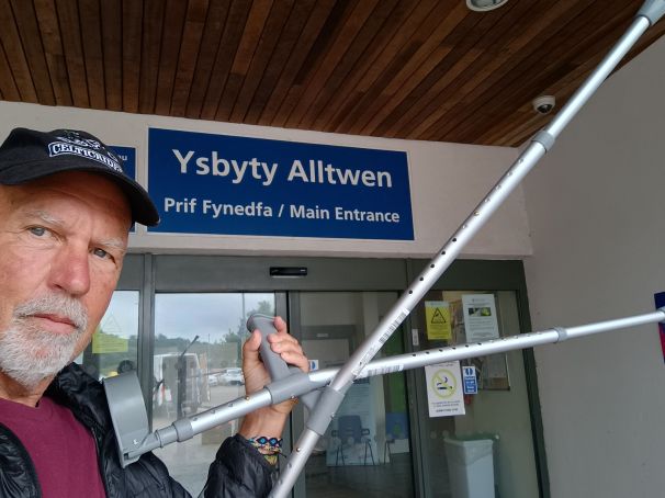 A person holding crutches in front of the main entrance to Ysbyty Alltwen hospital, with a wooden ceiling and a blue sign in English and Welsh.