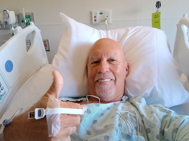 A smiling man in a hospital bed giving a thumbs up, dressed in a hospital gown with an IV attached to his arm.